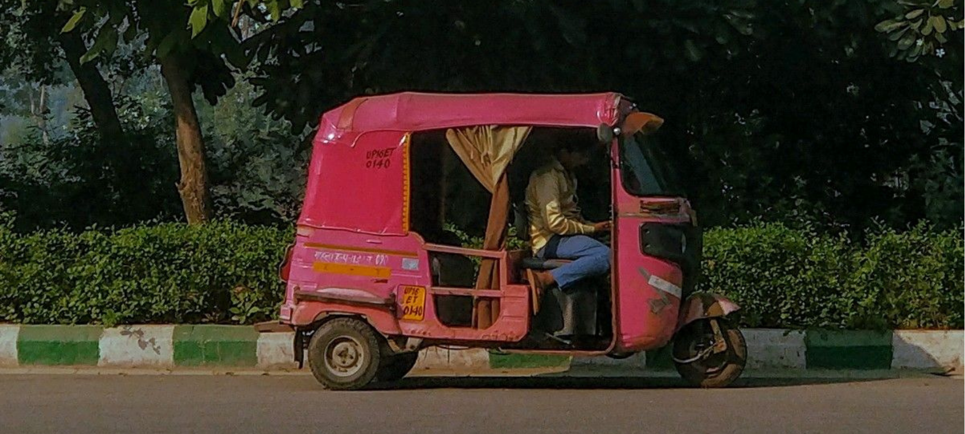 A pink three-wheeled auto rickshaw with a driver seated inside, photographed outdoors on a road with trees and hedges in the background. Large white text across the top reads 'YOUR ORDER IS ON ITS WAY!' and smaller text at the bottom reads 'beep beep'.