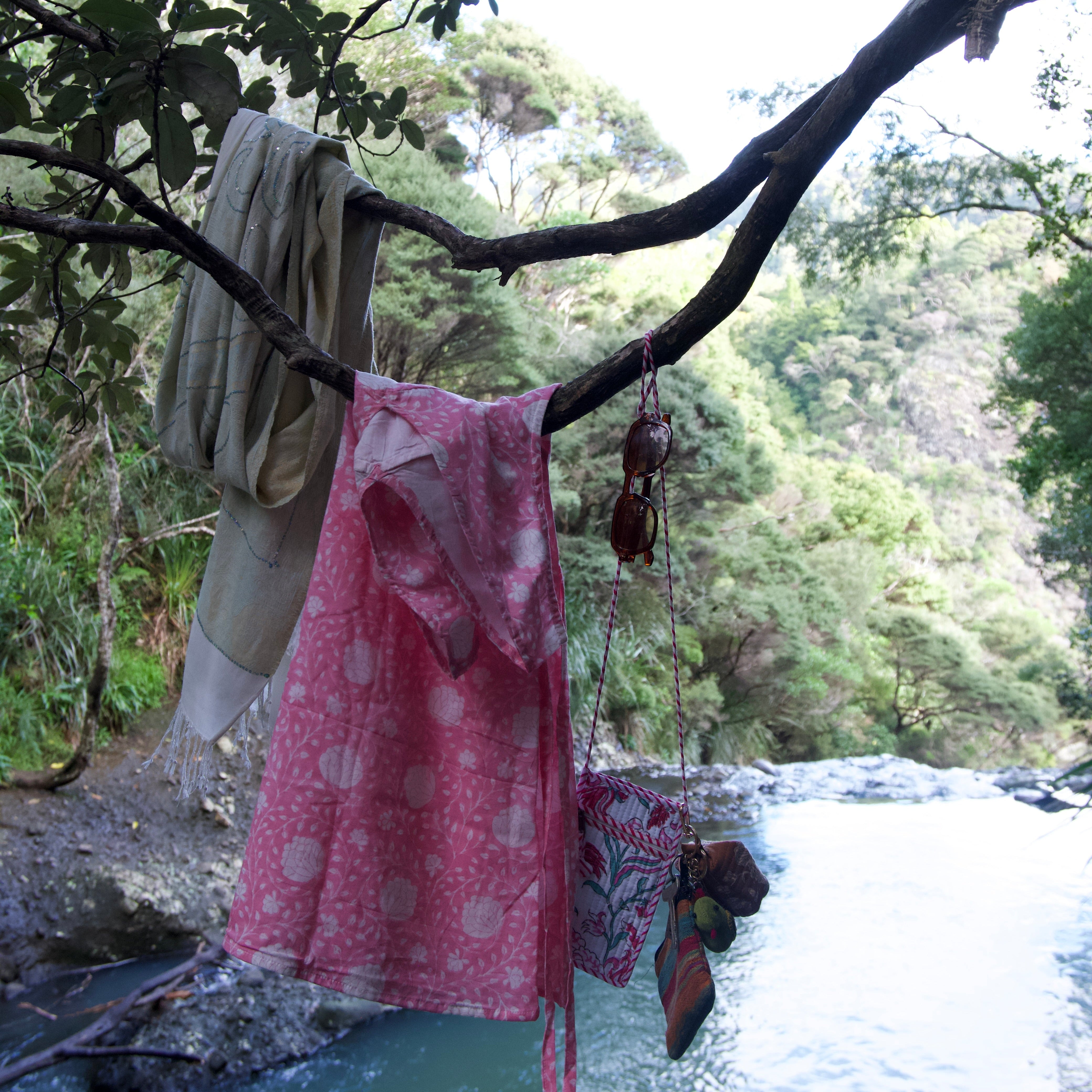 A pink patterned garment and other fabric items hang from tree branches over a stream in a natural outdoor setting with green forest in the background.